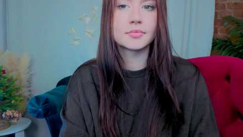 Video of a young woman with fair skin and long, straight brown hair, wearing a dark green, sheer, high-neck top, sitting on a plush red armchair in a room with light blue walls and a brick accent wall.