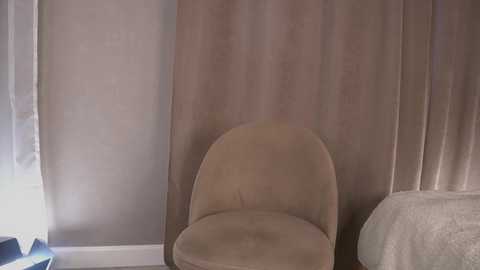 Video of a beige, upholstered chair against a beige curtain in a dimly lit room. The chair's curved back and smooth texture contrast with the textured curtain. A white bed with a fluffy white comforter is partially visible on the right.