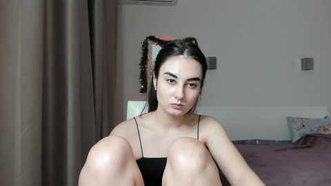 Video of a young woman with pale skin, dark hair, and cat ears, sitting on a bed in a dimly lit room with gray curtains.