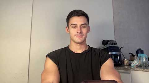 Video of a fit, light-skinned man with short dark hair, wearing a black sleeveless shirt, standing in a modern kitchen with granite countertops and appliances, including a kettle and a coffee machine.