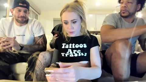 Video of a blonde woman with tattoos, wearing a black T-shirt with \"Tattoos Are Forever\" text, sitting on a couch surrounded by two men in casual clothes in a modern living room.