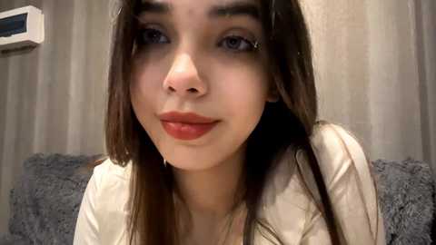 Video of a young woman with long brown hair, fair skin, and brown eyes, smiling softly. She wears a white top. Background includes beige curtains and a grey textured pillow.