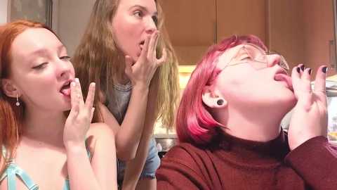Video of three women with fair skin, one with long blonde hair, one with red hair, and one with pink hair. They are laughing and making funny faces.
