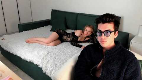 Video of a man and woman on a green couch with a white shag rug. The woman is in black lingerie, the man wears black glasses and a black jacket.