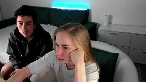 Video of a young couple in a modern living room: a man in a black jacket and a woman in a white shirt, both with long hair.