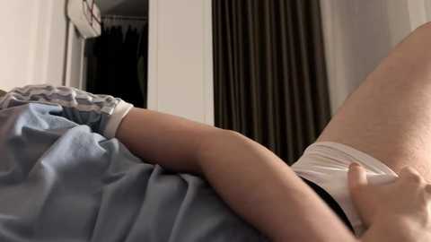 Video of a person's right arm in white shorts, lying on a bed with blue and white striped sheets, partially visible in a dimly lit room with a closed door and dark curtains.