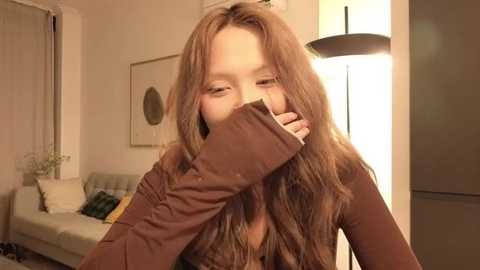 A video shows a young girl with long brown hair, covering her mouth with her hand, sitting indoors. She wears a brown sweater. Background includes a white couch, framed artwork, and a floor lamp.