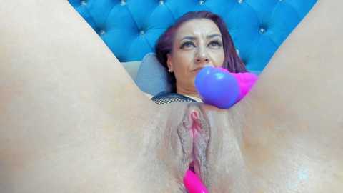 A video of a woman with fair skin, red hair, and light makeup, lying on a blue tufted headboard, with her legs spread, holding a purple dildo in her mouth.