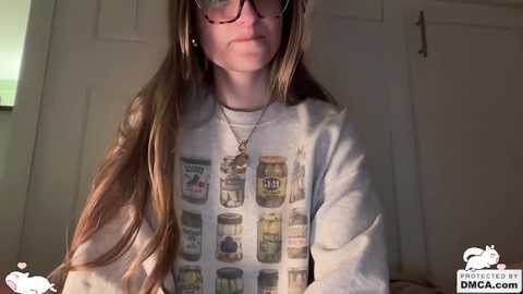 Video of a fair-skinned woman with long brown hair, wearing glasses, a gray sweatshirt with jars of honey, and a silver necklace. She stands indoors with a closed white door behind her.