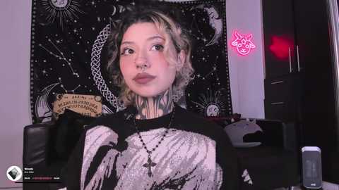 Video of a young woman with short, wavy blonde hair and pale skin, wearing a black and white graphic shirt. Background features a black tapestry with astrological symbols and a pink neon sign.