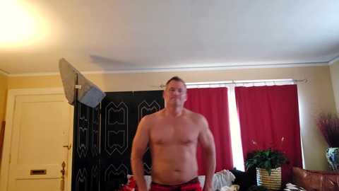 Video of a shirtless, muscular white man with short hair, wearing red shorts, standing in a room with a green screen, red curtains, and a potted plant.