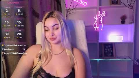 Media: Video of a blonde woman with fair skin and medium breasts in a black lace bra, seated on a white chair, with neon lights and a digital display in the background.