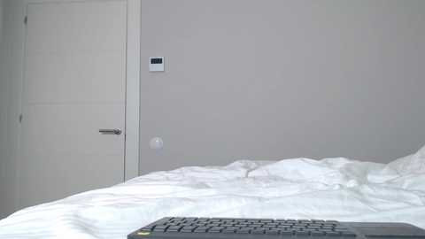 Video of a minimalist, modern bedroom with a white door, a thermostat, and a disheveled white bed. A black keyboard rests at the bottom edge.