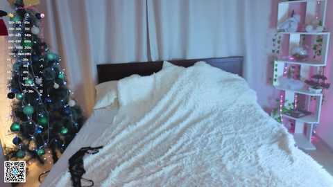 Video of a cozy bedroom with a Christmas tree decorated in green and silver, a white comforter covering a bed, and a pink shelving unit.