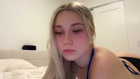 Media: Video of a young woman with fair skin and long, blonde hair lying on a bed. She has a neutral expression, wearing a blue bra and a choker. The room has white walls and a closed door.