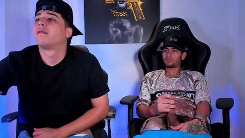 Video of two young men, one in a black cap and T-shirt, the other in a camo shirt, sitting in black gaming chairs in a brightly lit room with a poster on the wall.
