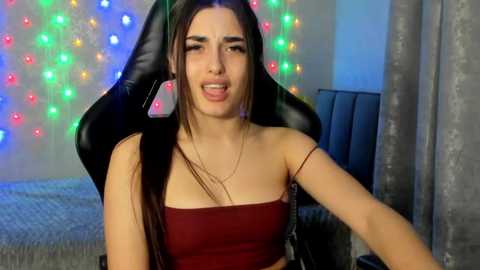 Video of a young woman with long dark hair, wearing a red strapless top, sitting on a black gaming chair with colorful string lights in the background.