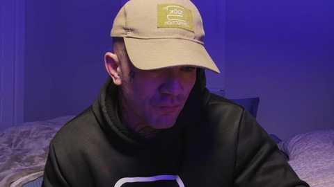 Video of a man with a light complexion wearing a beige baseball cap, black hoodie, and gray pillow in a dimly lit bedroom with purple lighting.