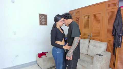 Video of a young Asian woman with glasses, medium build, wearing a black crop top and jeans, kissing a tattooed man in a black shirt, in a modern, beige-living room with wooden louvered doors.