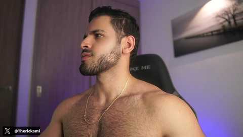 Video of a shirtless, bearded man with short, dark hair, wearing a gold chain necklace, sitting in a black gaming chair, with a minimalist room in the background.