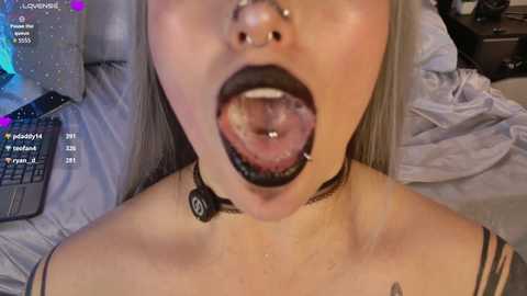 Video of a woman with pale skin, black lipstick, septum piercing, and a black choker, lying on a bed with a laptop displaying chat messages in the background.