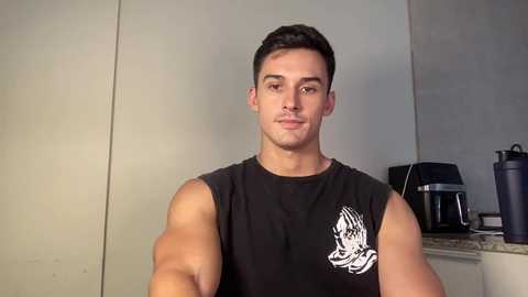 Video of a muscular young man with short dark hair, wearing a black sleeveless shirt with a white wolf graphic, standing in a modern kitchen with beige walls and a countertop holding a coffee maker.