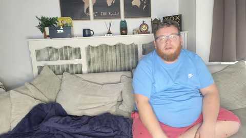 Video of a plus-size man with a beard and glasses, wearing a blue t-shirt and pink shorts, sitting on a beige couch with a blanket, surrounded by home decor in a cozy living room.