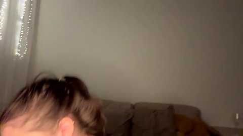 A video of a dimly lit room, featuring a young woman with brown hair in a ponytail, sitting on a gray couch against a plain white wall.