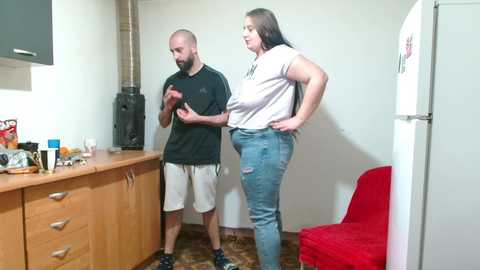 Video of a man with a beard in a black t-shirt and shorts, and a plus-sized woman in a white shirt and ripped jeans, standing in a small kitchen with wooden cabinets, a red chair, and a white fridge.