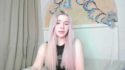 Video of a young Caucasian woman with long, pastel pink hair, wearing a black tank top, standing indoors with beige curtains and a wall-mounted blue floral arrangement.