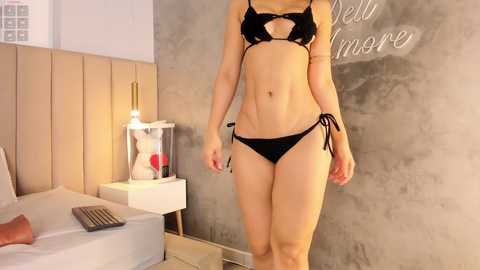 Video of a slim, fair-skinned woman in a black bikini with side-tie bottoms, standing in a modern bedroom with a wooden headboard, beige walls, and a \"Bella Is More\" chalkboard sign.