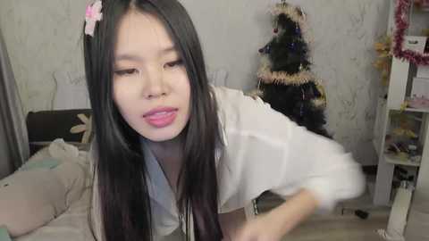 Video of an East Asian woman with long black hair, wearing a white jacket, making a funny face in a cozy, Christmas-decorated room with a tree.
