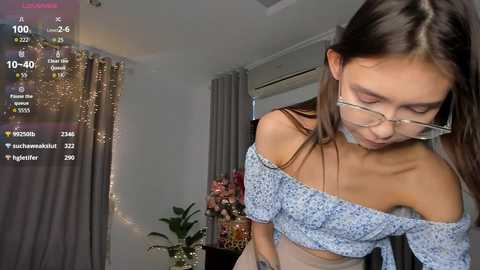 Video of an Asian woman with glasses, wearing a light blue off-shoulder top, standing in a room with a potted plant, curtains, and a digital overlay showing social media metrics.