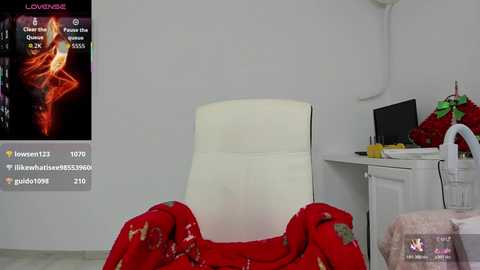 Video of a cozy, minimalist room with a white, cushioned chair draped in a red, floral blanket. In the background, a white desk holds a computer and Christmas decorations. A pop-up window displays a steamy, red-themed video with a woman in lingerie.