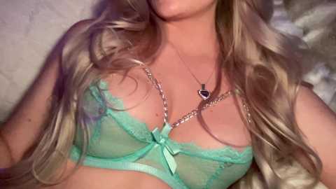 Video of a light-skinned woman with long, wavy blonde hair, wearing a sheer, mint-green lace bra with a bow, silver chain necklace, lying on a soft, white bedspread.
