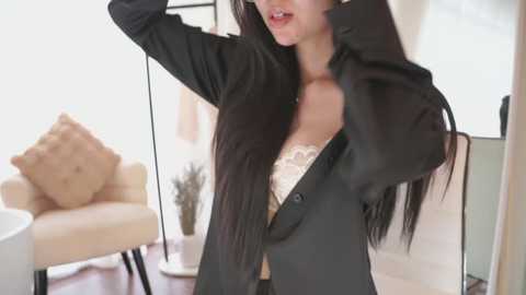 Video of an Asian woman with long black hair, wearing a black blazer and white lace bra, adjusting her hair in a modern living room with beige furniture and a potted plant.
