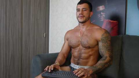 Video of a muscular, tattooed man with short dark hair, wearing gray boxer briefs, seated on a gray couch, typing on a keyboard, with a framed picture in the background.