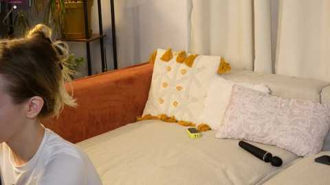 Video of a woman with blonde hair in a bun, sitting on a beige couch with an orange armrest, adorned with a patterned pillow and a yellow remote.