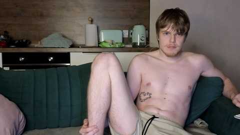 Video of a shirtless, fair-skinned young man with light brown hair, sitting on a green couch in a modest kitchen. He wears beige shorts and has a tattoo on his left side.