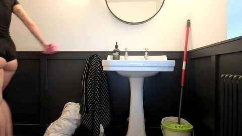 Video of a person in a black top and white shorts cleaning a white sink in a black-paneled bathroom with a striped towel and a red broom nearby.