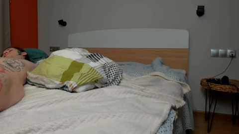 A video of a man, tattooed on his chest, sleeping in a bed with a white duvet, surrounded by a messy room with a wooden headboard, gray walls, and a wicker nightstand.