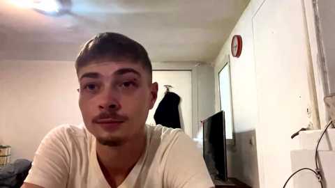Video of a young man with short brown hair, wearing a white T-shirt, indoors. Background shows a white wall, a television, and a person standing in the doorway.