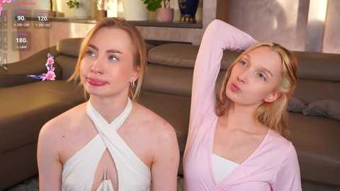 A video of two young women in a cozy living room. One woman, with fair skin and blonde hair, wears a white halter top; the other, also fair-skinned, wears a pink cardigan.