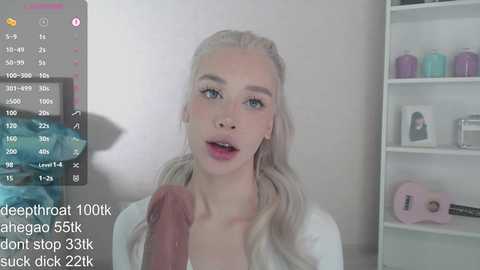 Video of a young, blonde, fair-skinned woman with blue eyes, wearing a white top, performing oral sex on a large, erect penis in a brightly lit room.