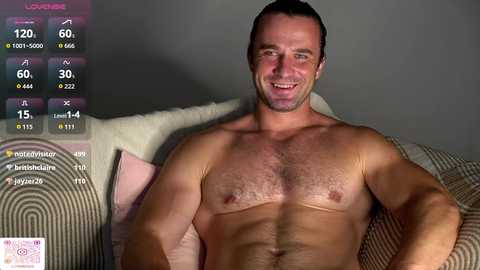 Video of a shirtless, muscular, white man with short dark hair, smiling, lying on a couch with a striped blanket. Background includes a gray wall and a digital display with fitness stats.
