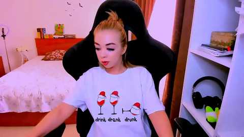 Video of a blonde woman with light skin and medium build, wearing a white t-shirt with a cartoon design of two wine glasses, sitting on a black gaming chair in a modern bedroom.