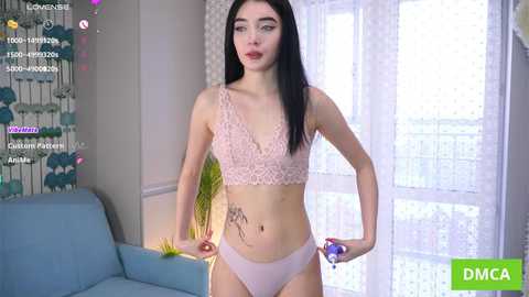 Video of a slender, fair-skinned woman with long black hair, wearing pastel pink lace lingerie. She stands confidently in a modern living room with white curtains and a blue couch.
