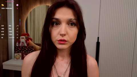 Video of a young woman with long, dark brown hair, wearing a sleeveless top, standing in a bedroom with a white wardrobe and a mirror.