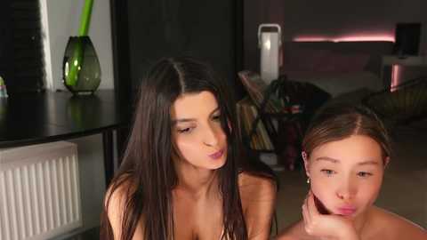 Video of two young women, one with long black hair, another with brown hair, sitting close, in a modern, dimly-lit room with a black table, green vase, and bookshelf.