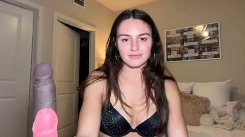 Video of a young woman with long brown hair, fair skin, and a medium build, wearing a black bra, holding a large pink dildo, in a bedroom with beige walls, a white bed, and a framed picture.